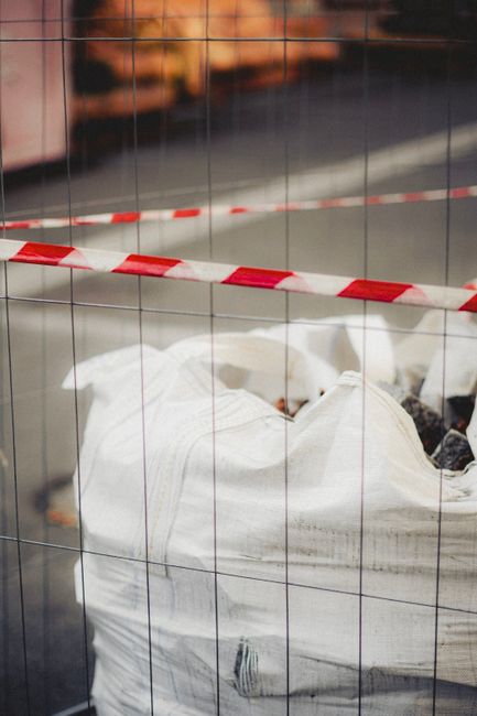 The image shows a large white sack filled with dark waste materials, situated on a paved surface which appears to be part of a driveway or outdoor area. The sack is made of a woven, sturdy fabric with